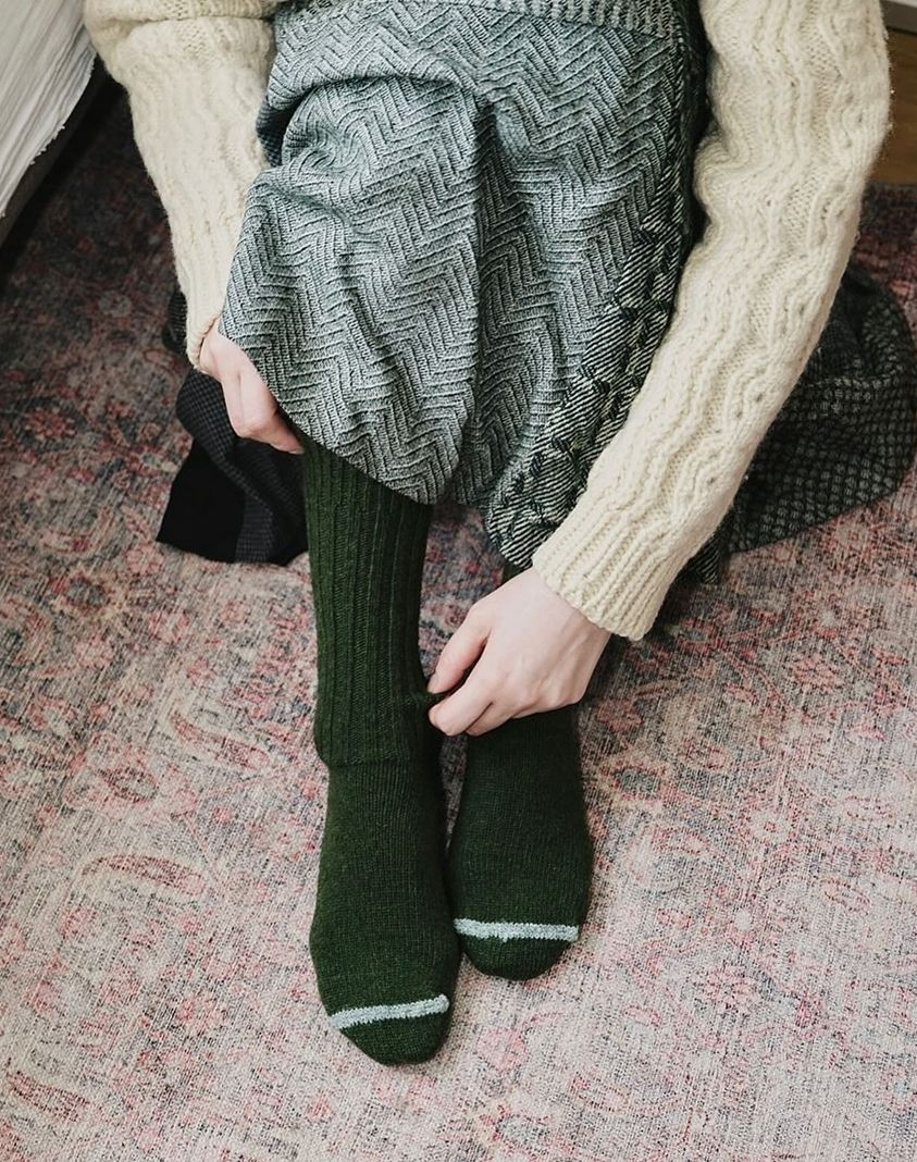 An image of a woman wearing Khaki socks from Nishiguchi Kutsushita,, she is wearing a cream cable knit weather and skirt sat on the floor.
