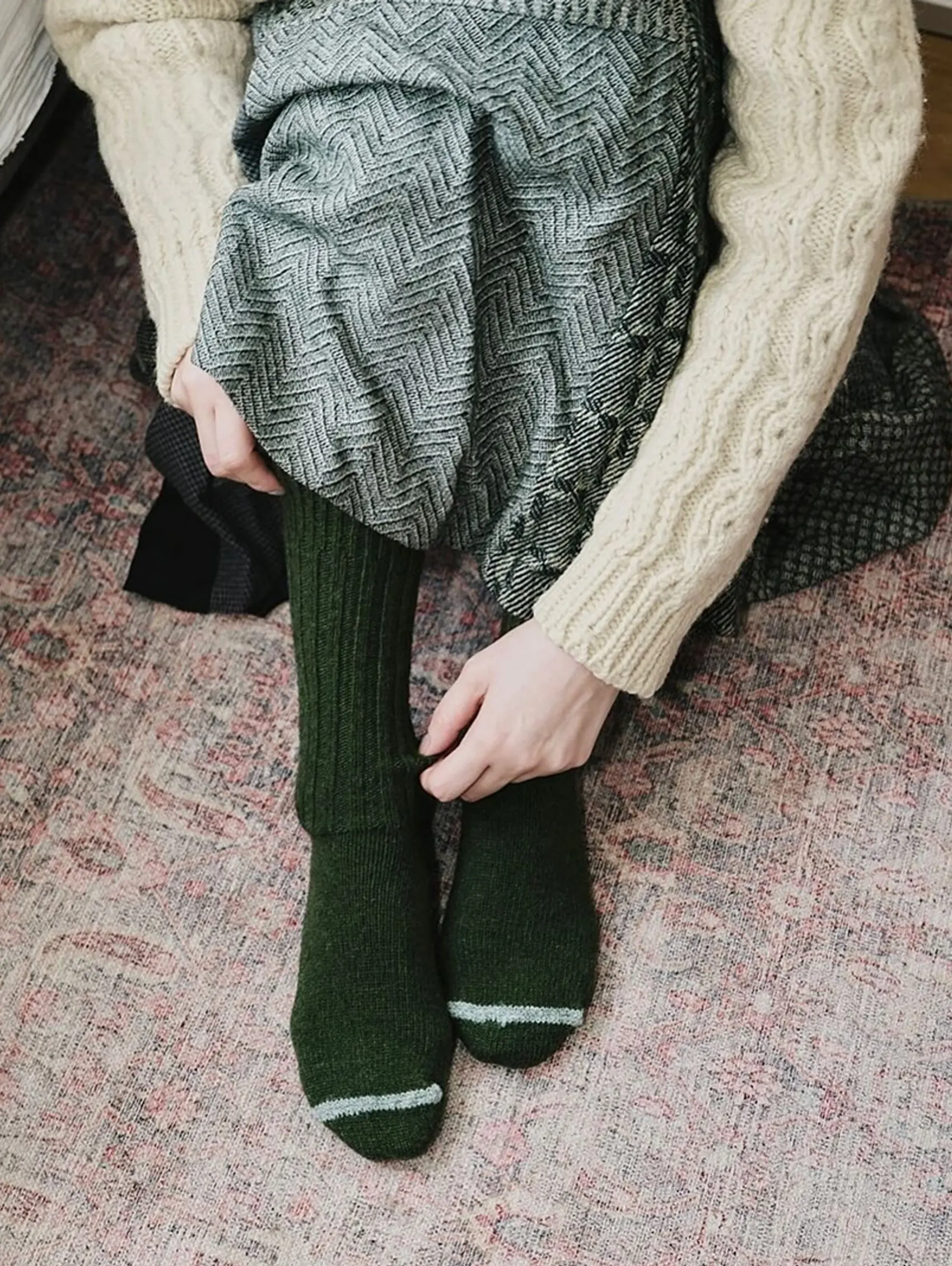 An image of a woman wearing Khaki socks from Nishiguchi Kutsushita,, she is wearing a cream cable knit weather and skirt sat on the floor.