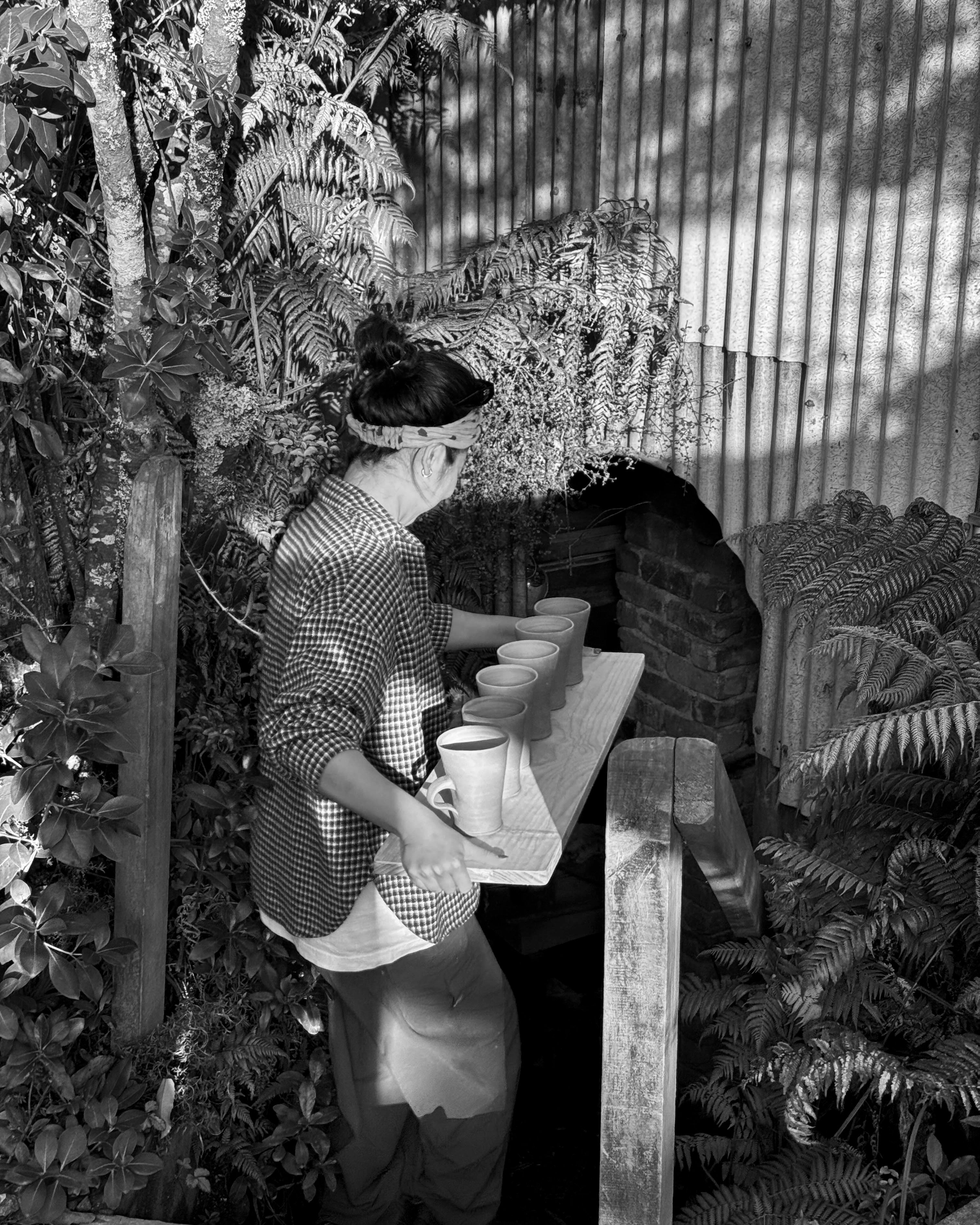 Kaiqi a ceramic artist, holding a tray of ceramic cups going into a bisque firing, the tin shed is surrounded by nature and native New Zealand trees