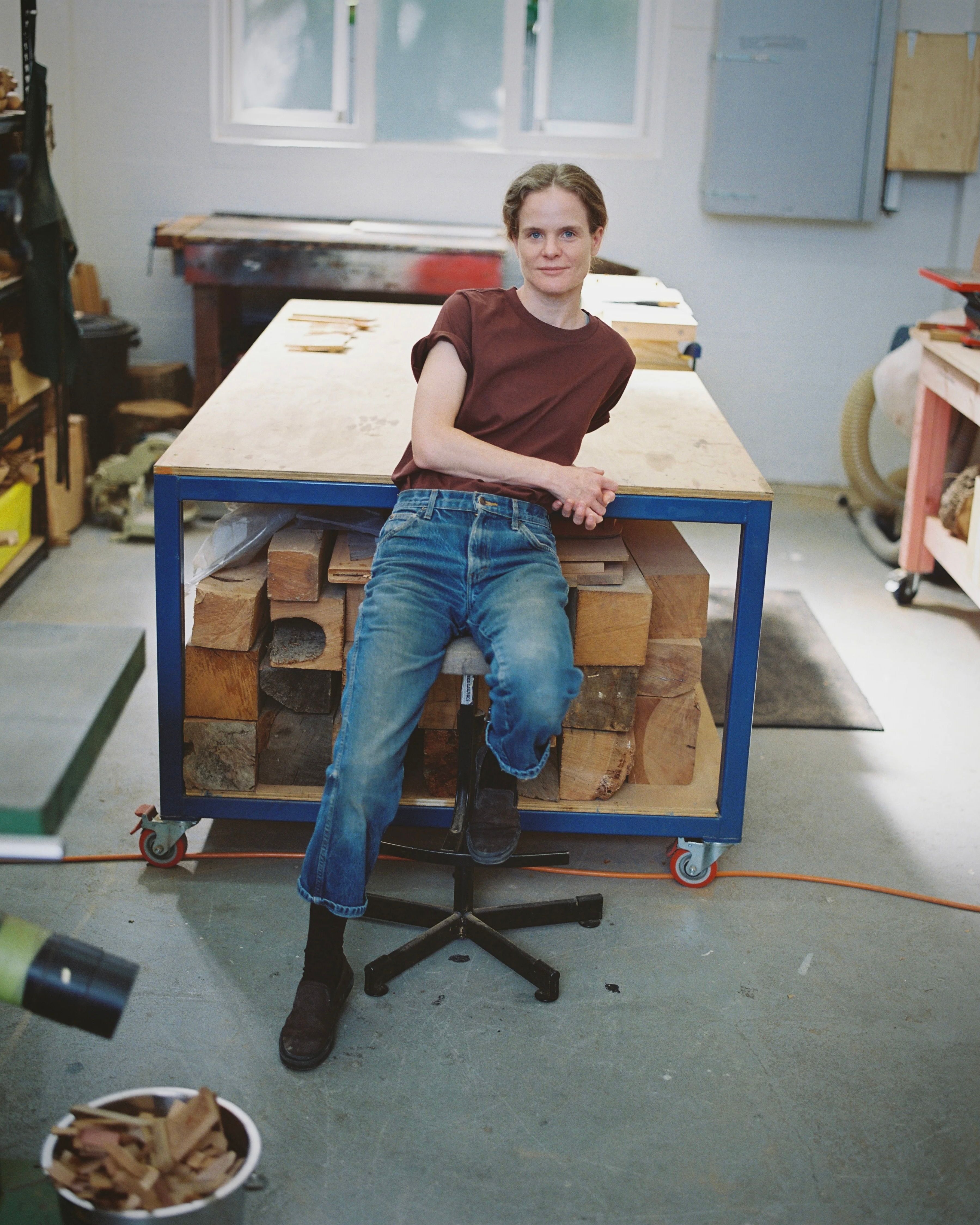A portrait image of New Zealand woodworker, Courtney Petley in her workroom with a large table behind her.