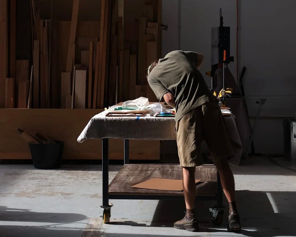 An image of Emile Drescher a woodworker from Auckland New Zealand, in his workshop with planks of wood stacked up on the walls, he is leaning over a workshop table