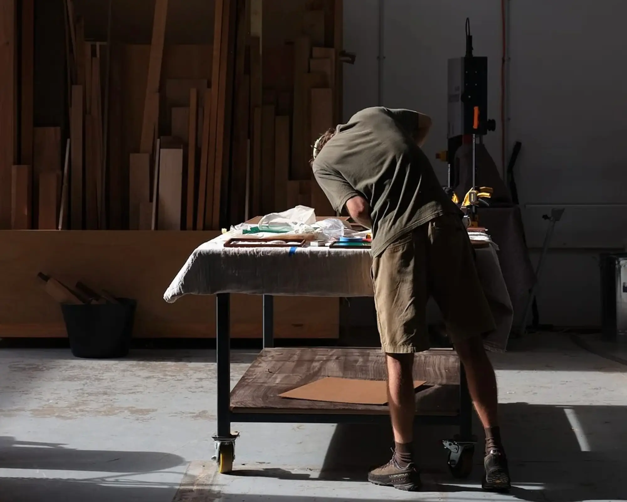 An image of Emile Drescher a woodworker from Auckland New Zealand, in his workshop with planks of wood stacked up on the walls, he is leaning over a workshop table