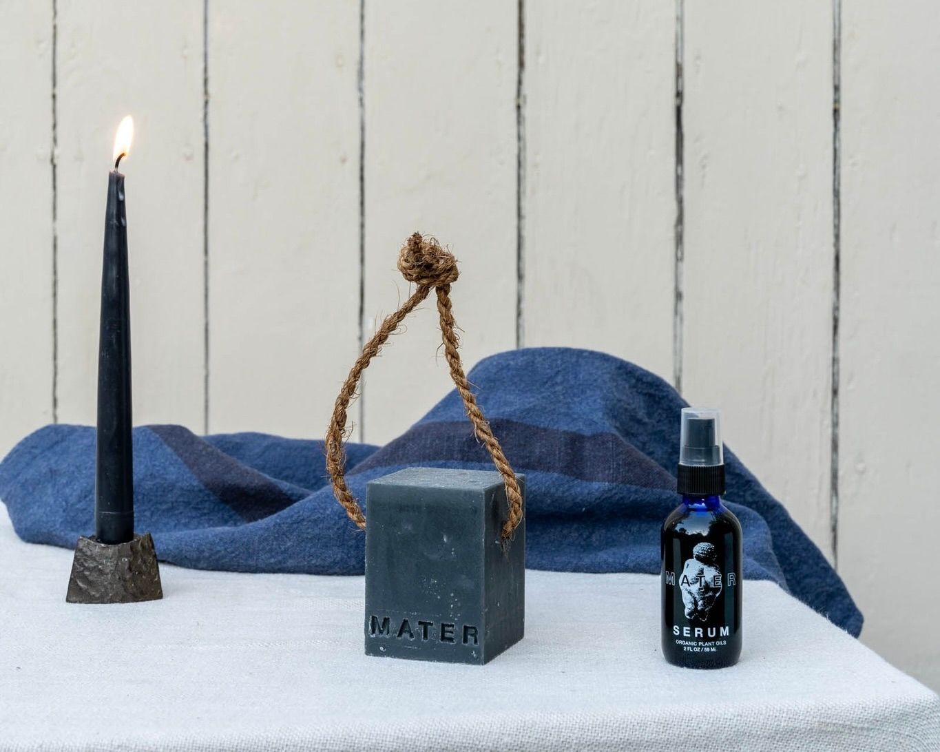 An image from Mater soap, their organic facial and hair serum made from natural plant oils alongside a block of cold pressed soap with a navy candle on a table