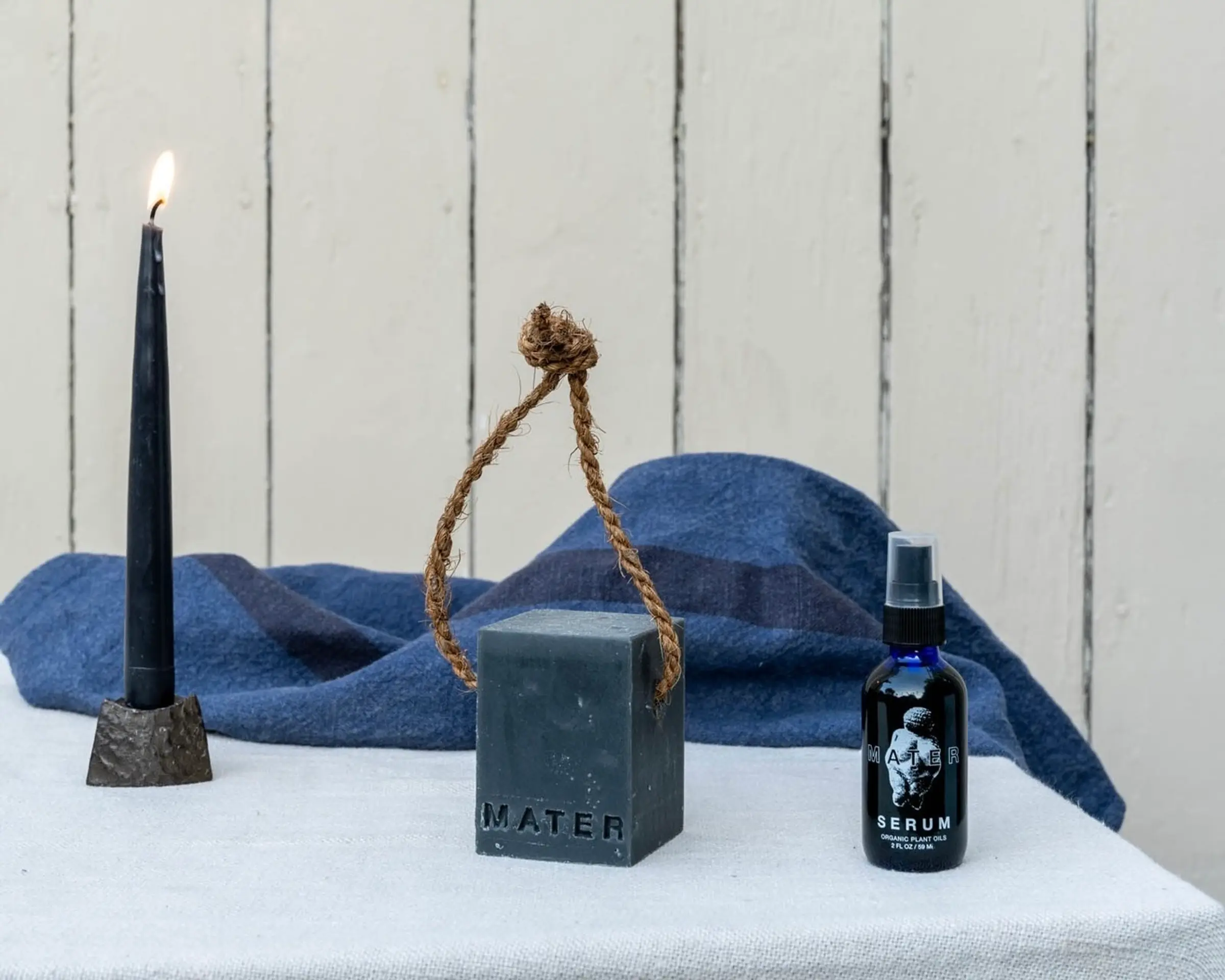 An image from Mater soap, their organic facial and hair serum made from natural plant oils alongside a block of cold pressed soap with a navy candle on a table