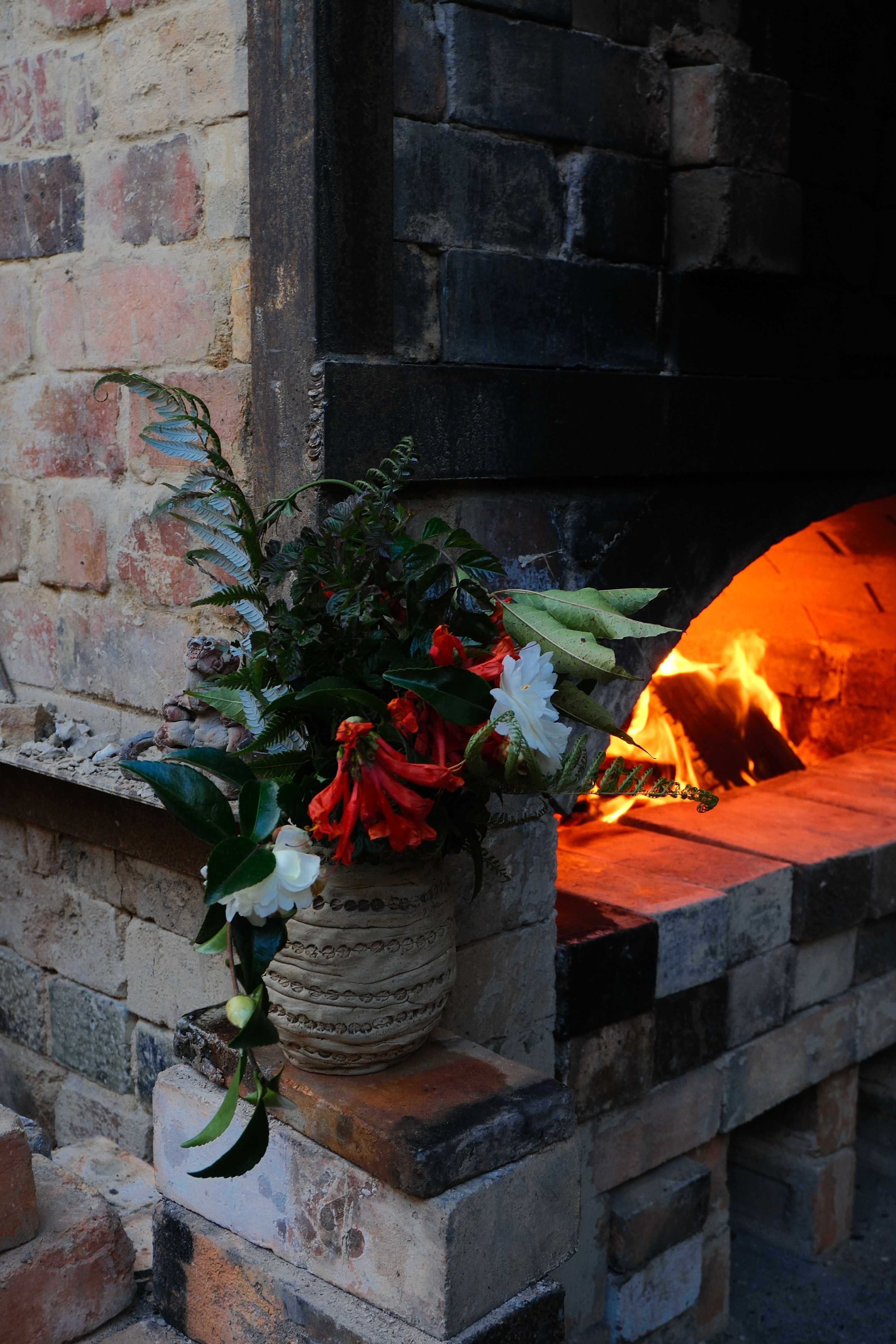 A wood fired kiln made from bricks with a ceramic vase filled wth flowers and a fire burning behind it at Driving creek pottery in the Coromandel of New Zealand