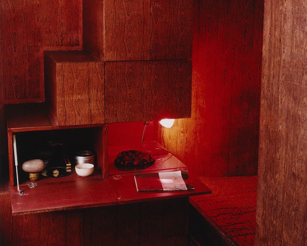 An image from Scenery magazine number 5 of a room with timber covered walls and red lacquer accents of a fold out table side bench with ceramics a light and a book