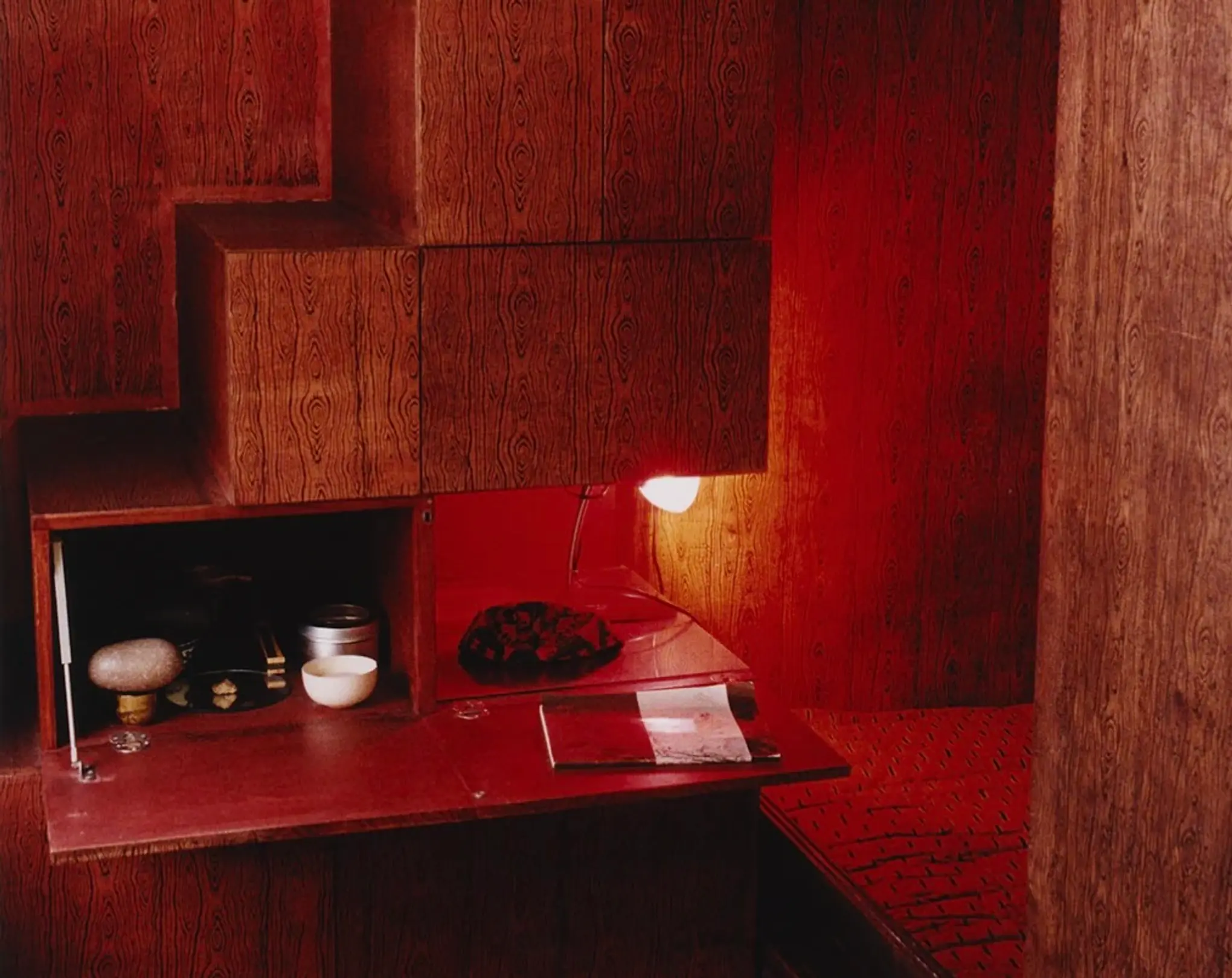 An image from Scenery magazine number 5 of a room with timber covered walls and red lacquer accents of a fold out table side bench with ceramics a light and a book