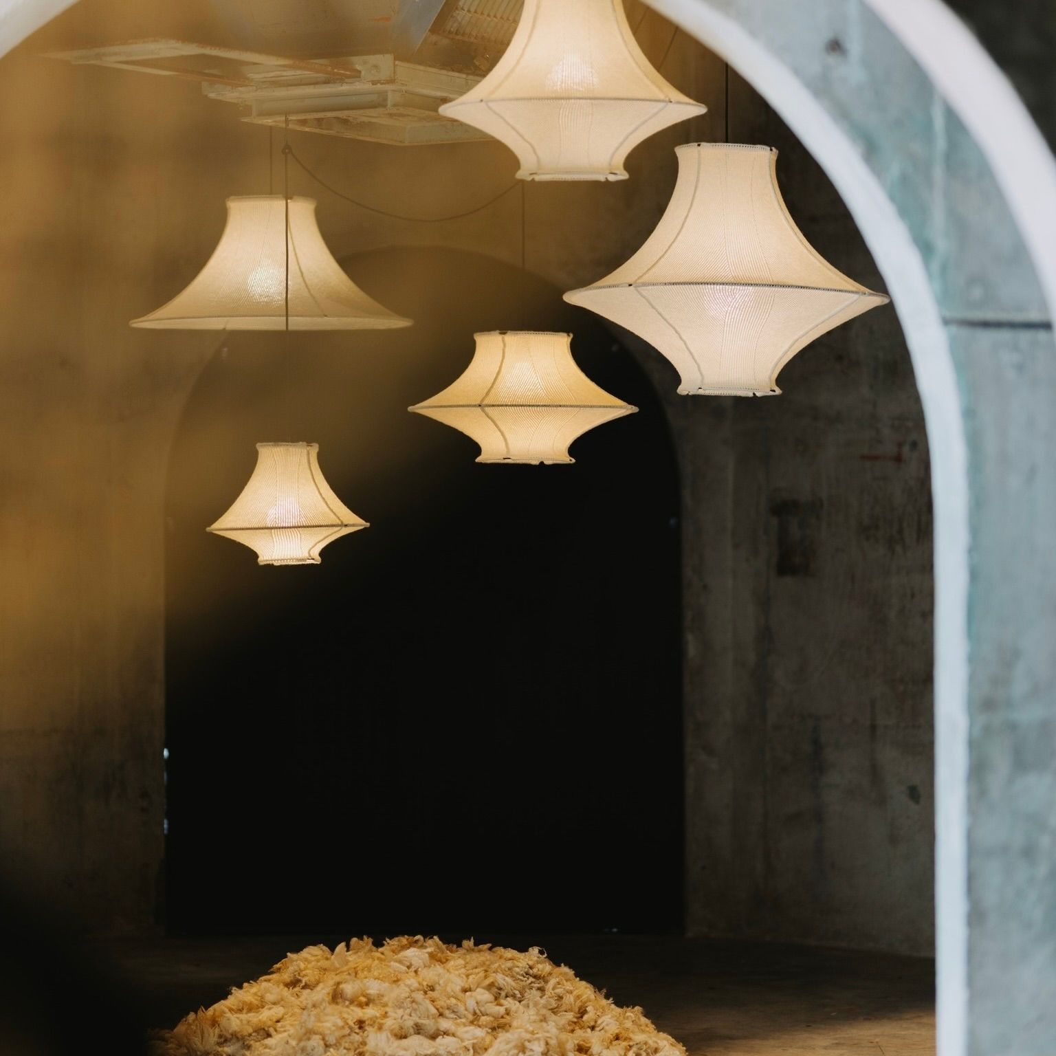 LOF HIWA wool pendant lights illuminating  a dimly lit concrete room with softly diffused lighting . A pile of natural New Zealand wool sits on the floor below, framed by a large archway in the foreground.