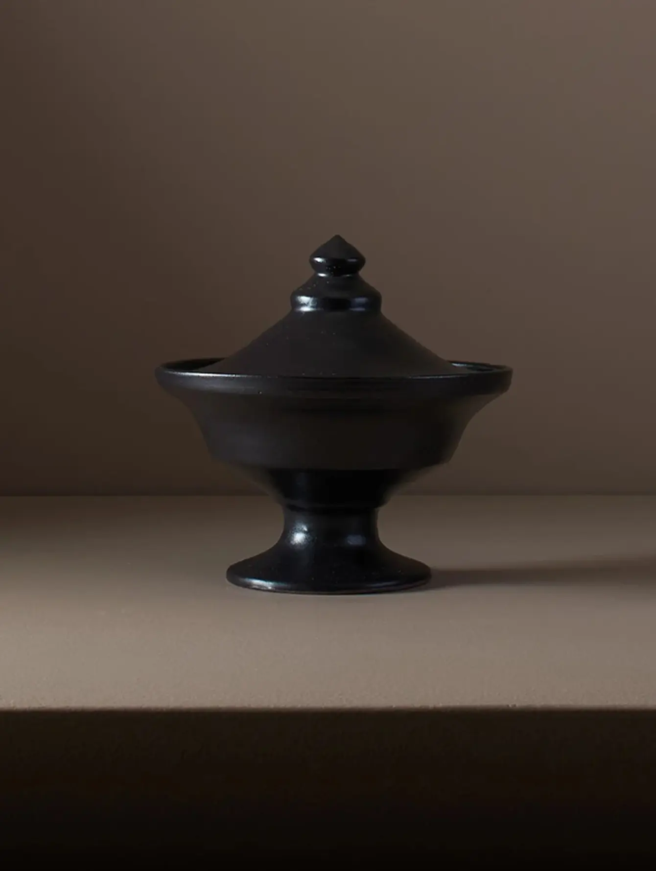 A ceramic Black lidded vessel from Kirsten Druburgh pottery with a pointed lid, shape inspired from Etruscan pottery, sitting on a brown background