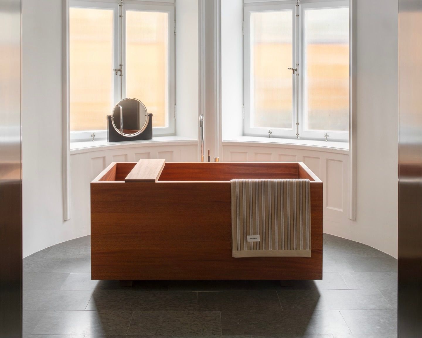 A magniberg beige stripe towel hung over a wooden bath tub in a light filled bathroom with a small circle mirror sitting on behind it