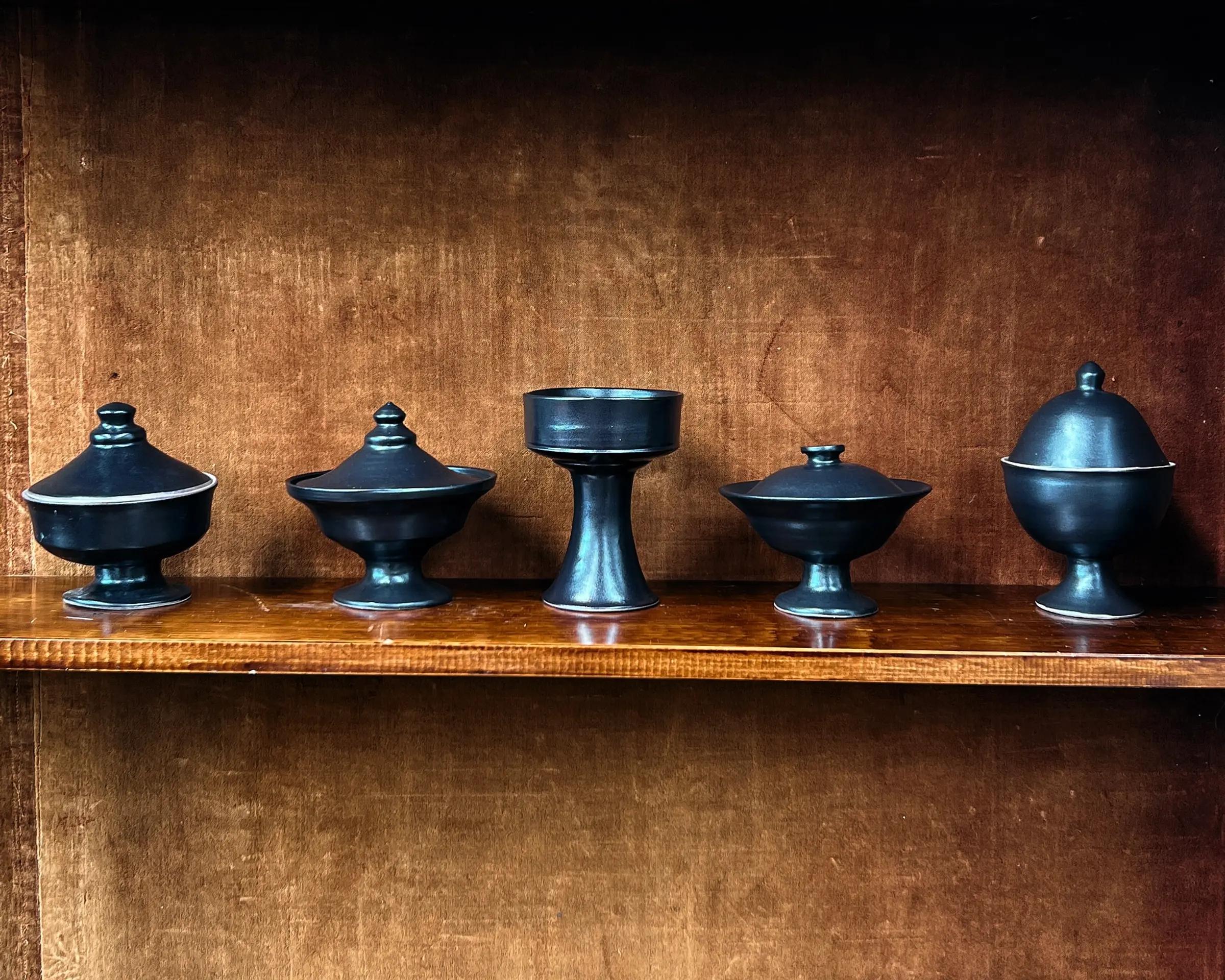 Hand-thrown black stoneware lidded vessels by Kirsten Dryburgh in a vintage wooden cabinet with a brown velvet background 