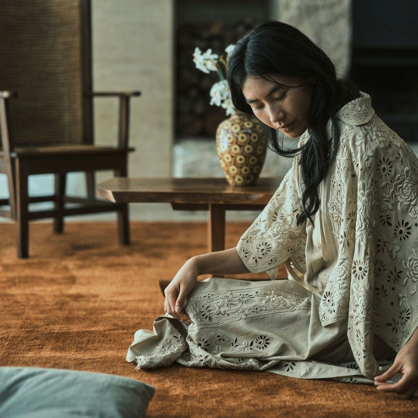 an inset shot of a model on a silk and bamboo rug with vintage chair and table in the background with a vase of flowers