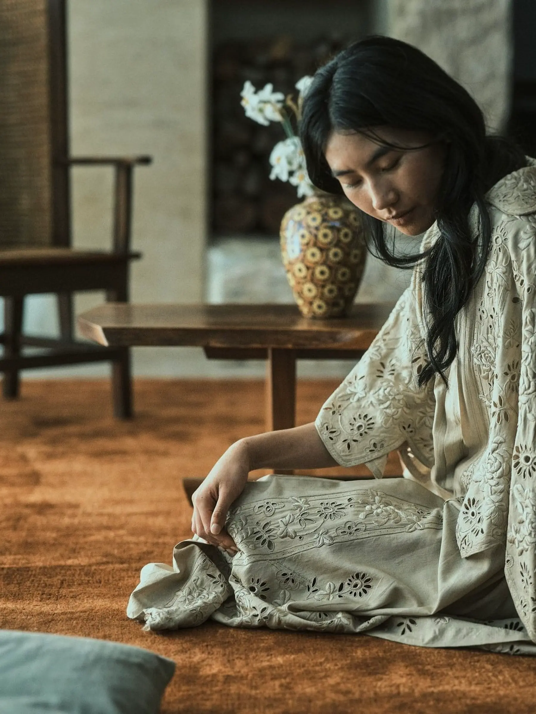 an inset shot of a model on a silk and bamboo rug with vintage chair and table in the background with a vase of flowers