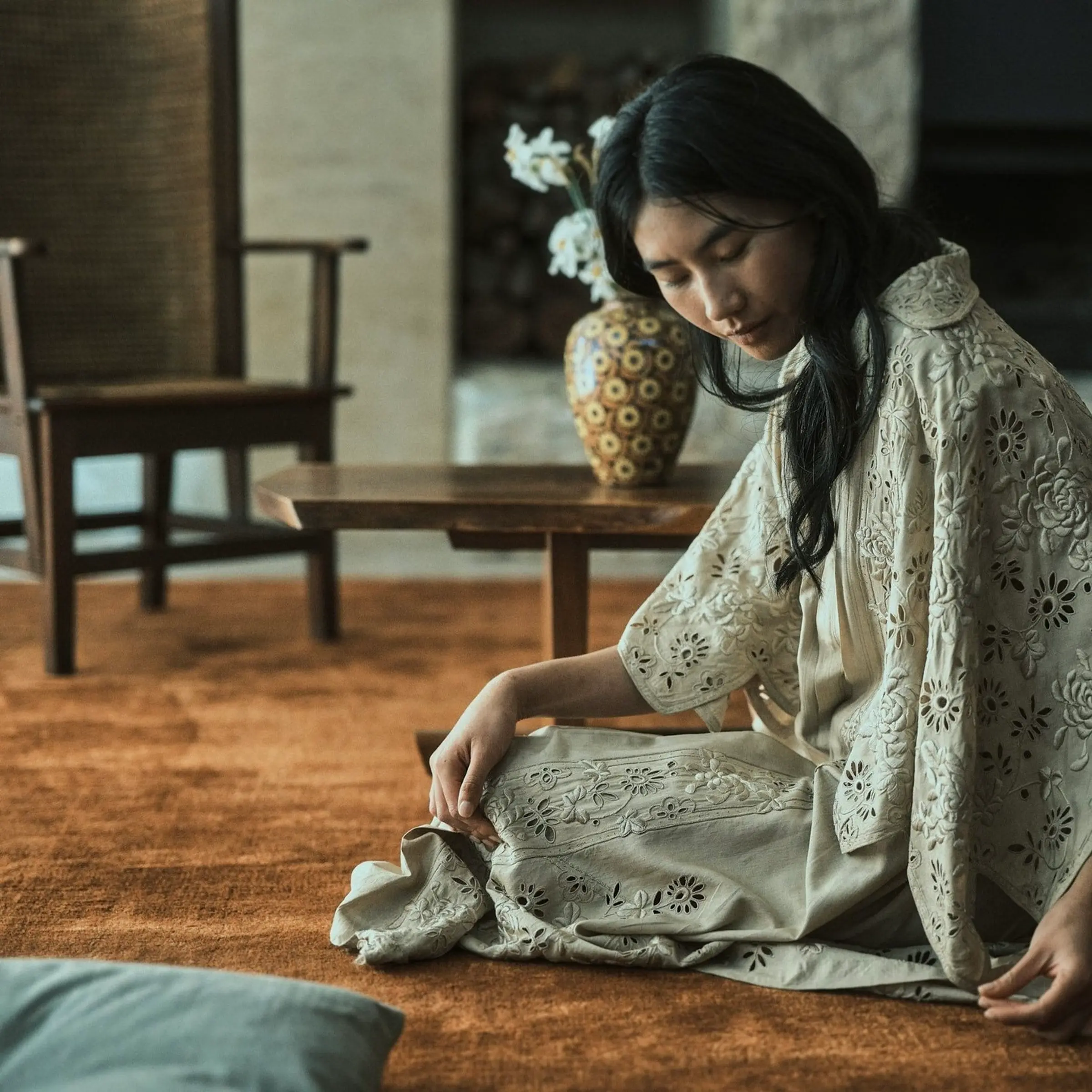 an inset shot of a model on a silk and bamboo rug with vintage chair and table in the background with a vase of flowers
