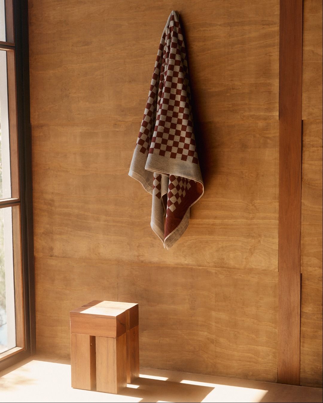 A checkered bath towel hung on a wooden wall with a square wooden stool on the floor 