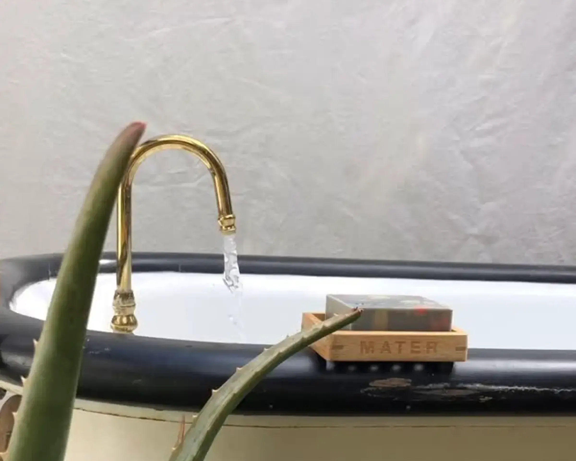 An image from Mater Soap with a bar of their natural cold pressed soap sitting on the side of a bath tub outside with a gold tap