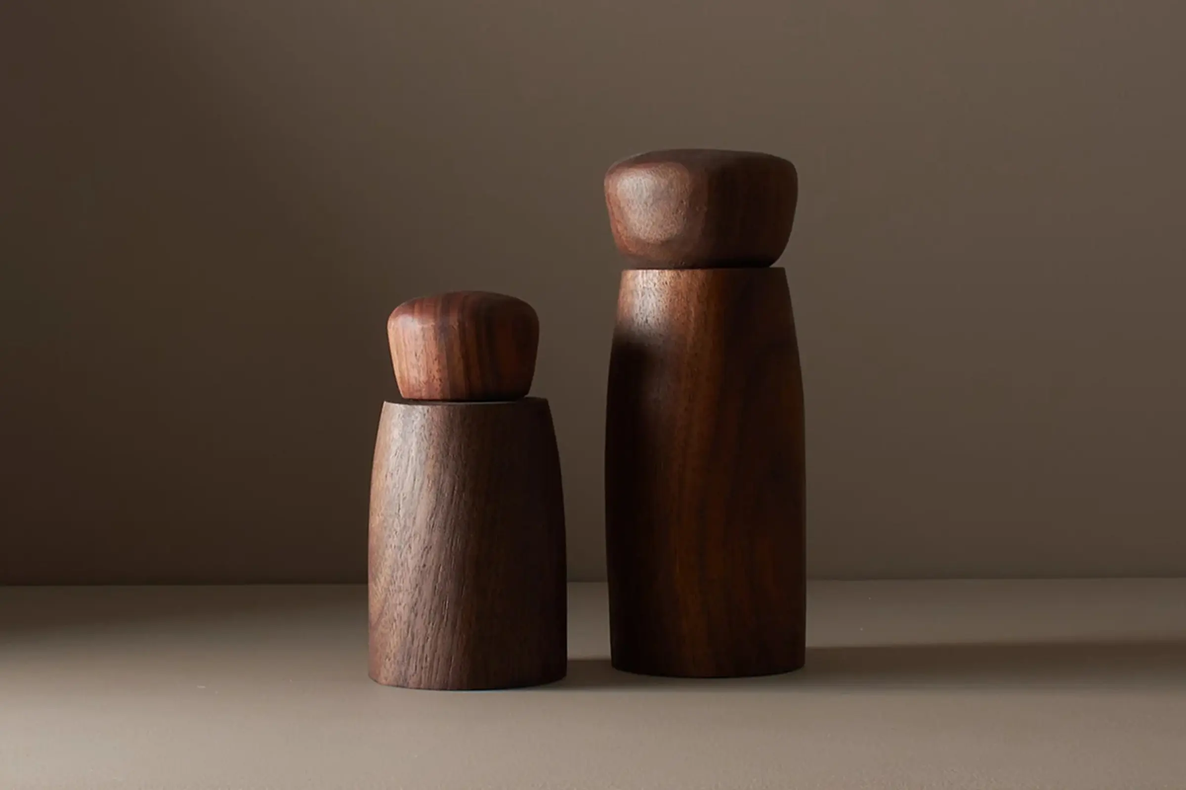 A pair of walnut wood salt and pepper mills in a deep chocolate colour by wood worker Elise McLauchlan from Canada sitting on a brown background