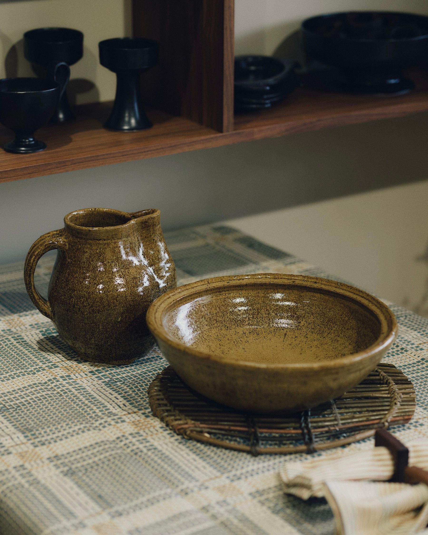 Dryburgh Pottery caramel green glazed harvest jug and large serving bowl on a tabletop with a Studio Ford block-printed tablecloth and woven willow trivet