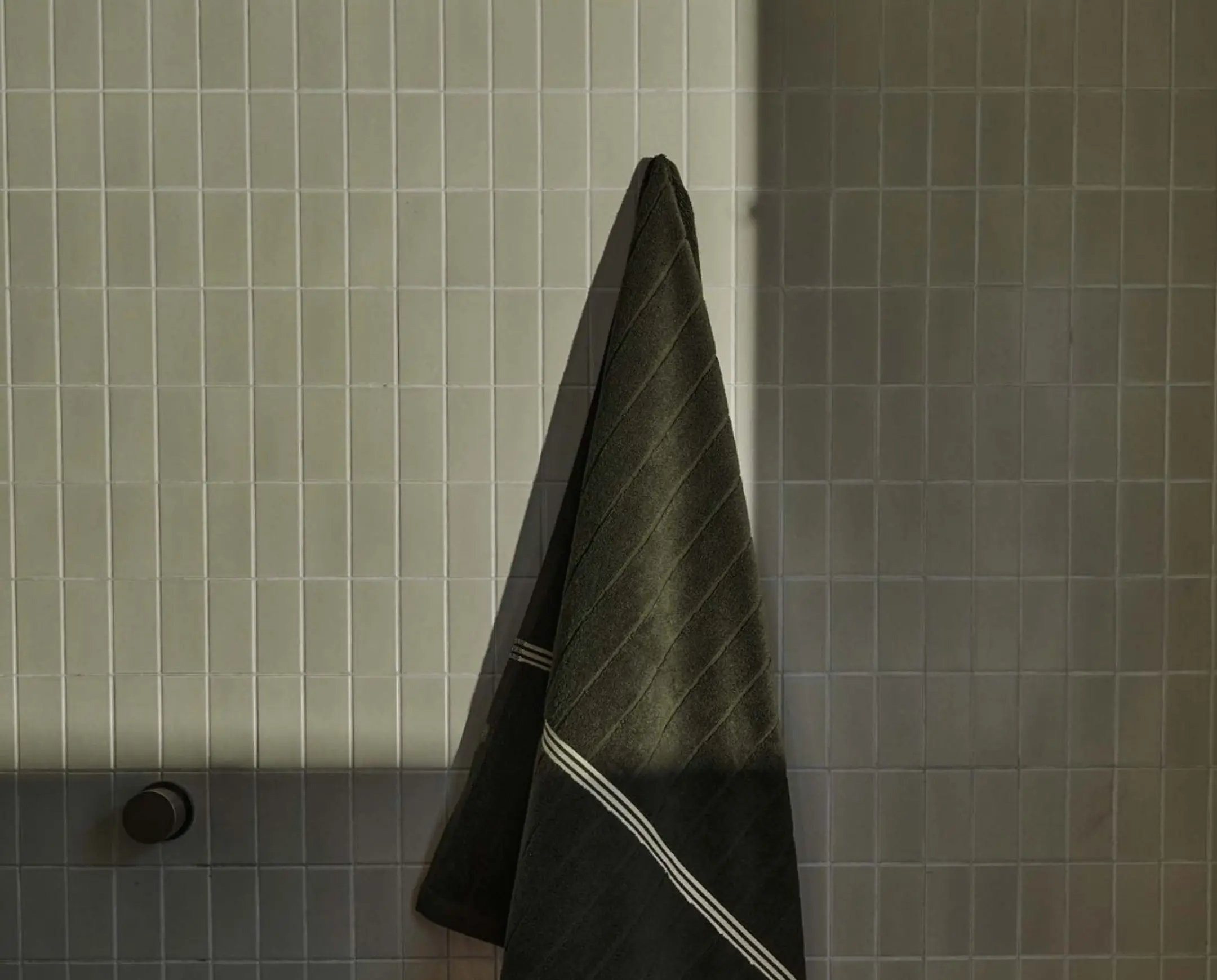 A deep green bath towel on a hook with pale green tiles behind it