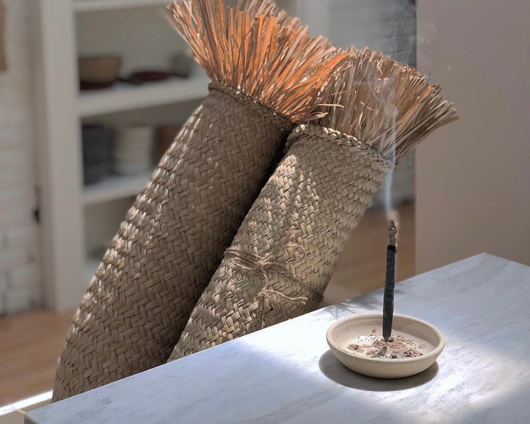 a palo Santo incense stick from Incausa burning on a tabletop with woven mats behind it