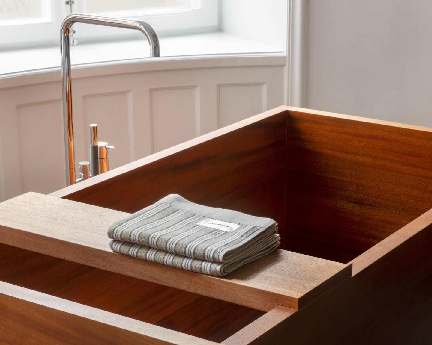A stack of Magniberg Wall Street hand towels in dark grey folded on a tray over a wooden bath tub in a cream walled home with a silver tap hanging over