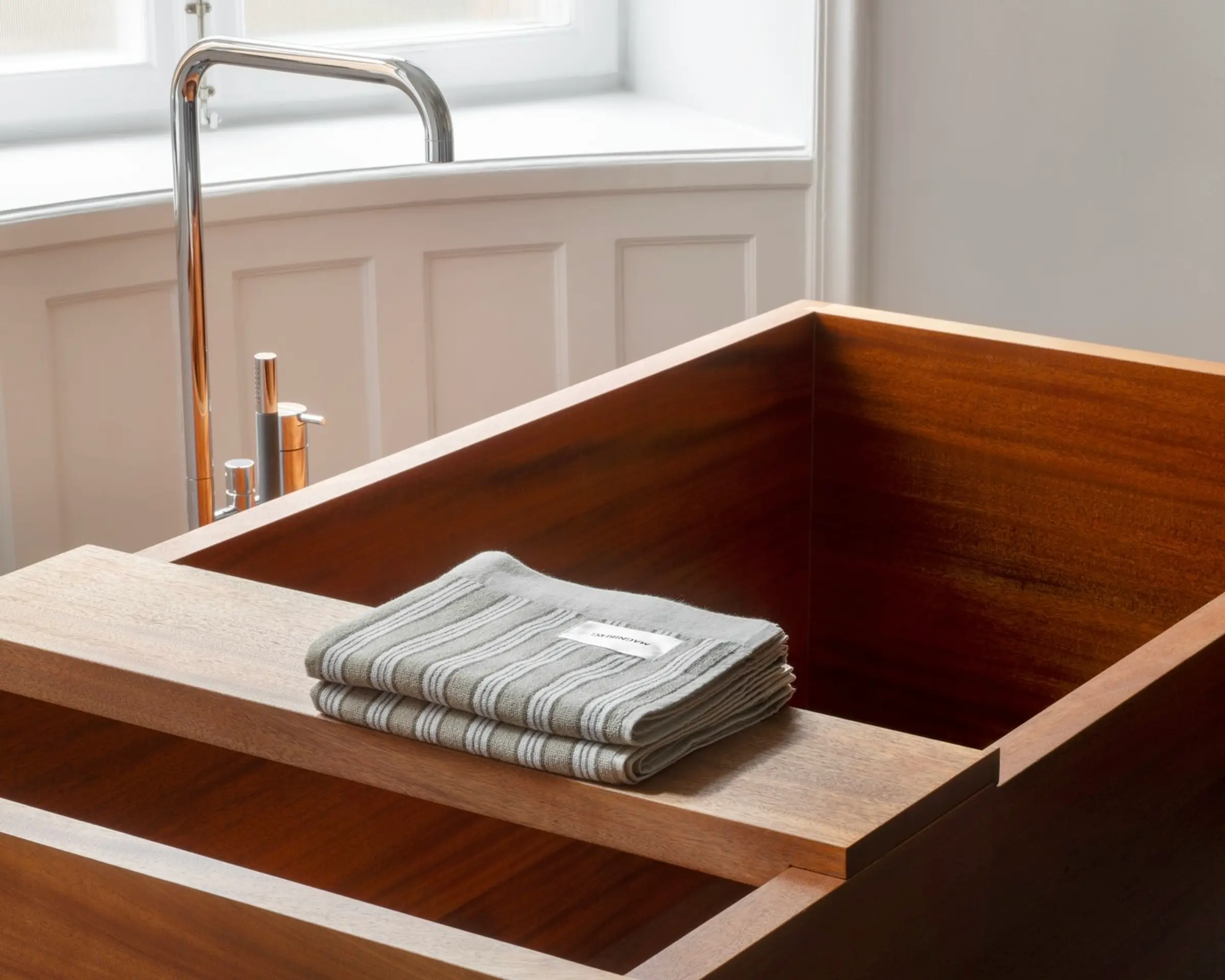 A stack of Magniberg Wall Street hand towels in dark grey folded on a tray over a wooden bath tub in a cream walled home with a silver tap hanging over