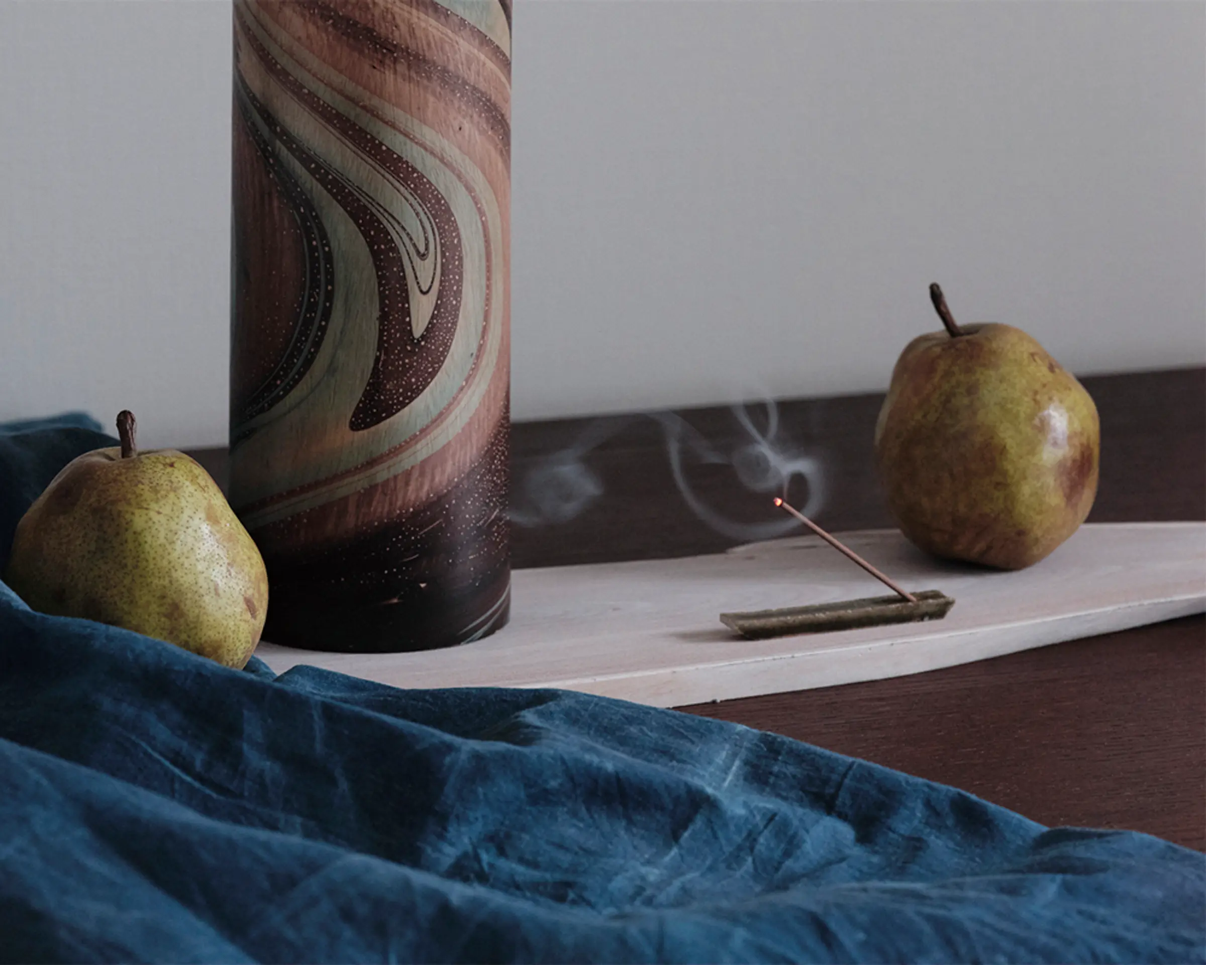 Burning incense on a desk next to 3 pears and a vase with a deep teal textile in the front