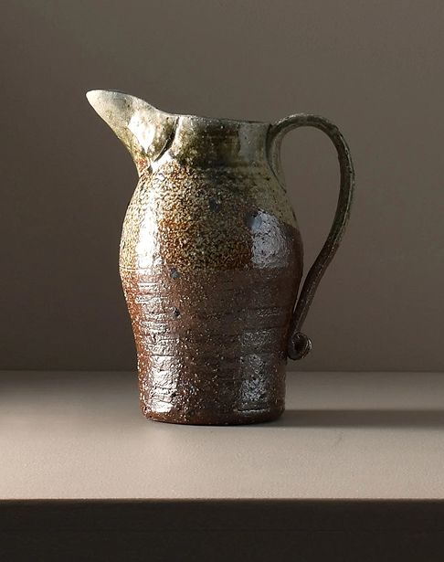 A wood fired ceramic jug created by Kaiqi Qiq, with tones of rust and sage along the glaze. Made in the Coromandel in New Zealand. Sitting on a brown background