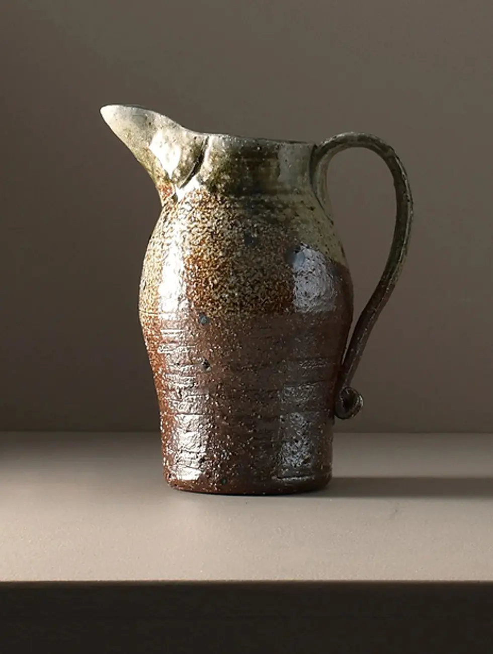 A wood fired ceramic jug created by Kaiqi Qiq, with tones of rust and sage along the glaze. Made in the Coromandel in New Zealand. Sitting on a brown background