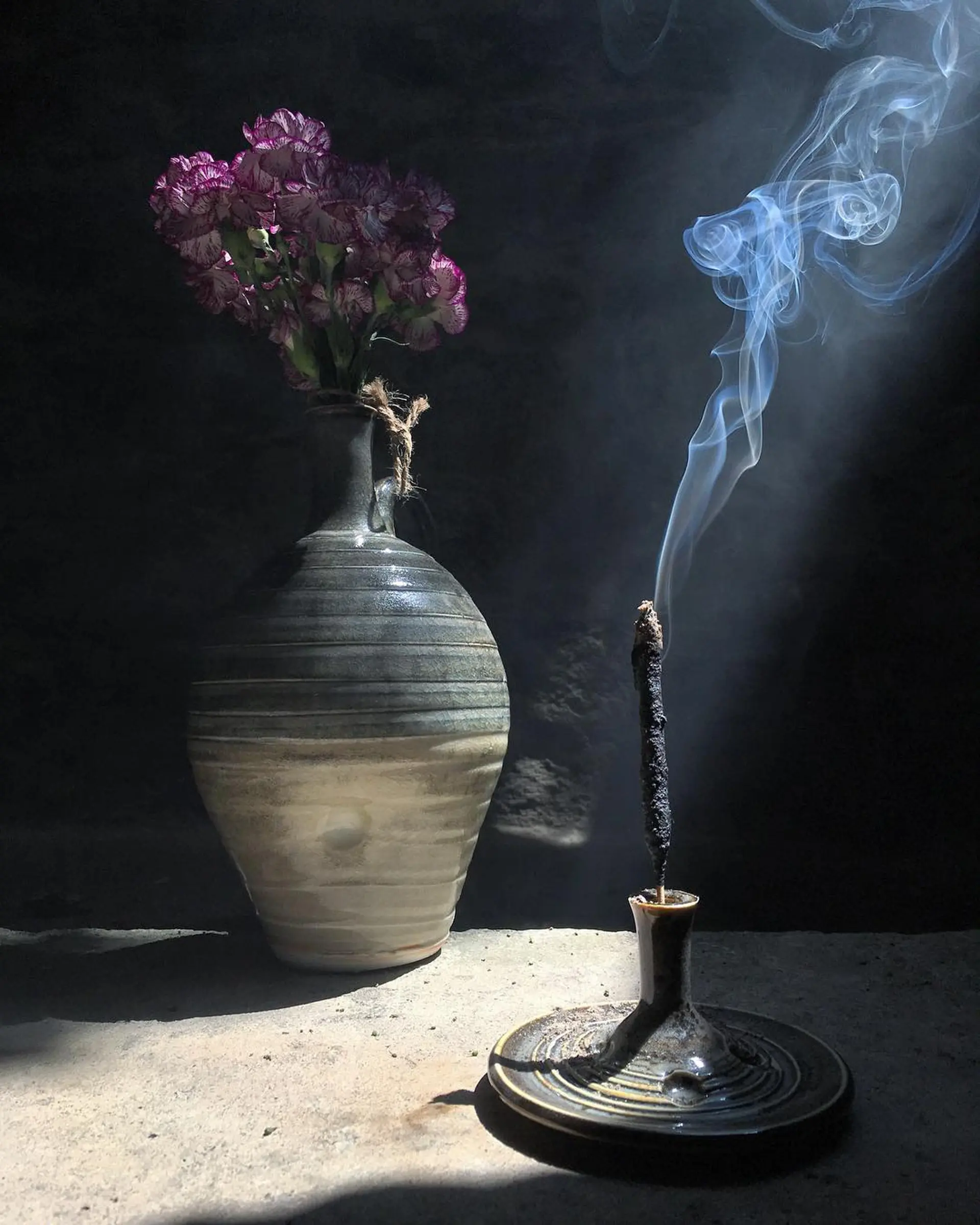 Incausa incense stick burning in a moody image with a pottery vase and flowers in the background