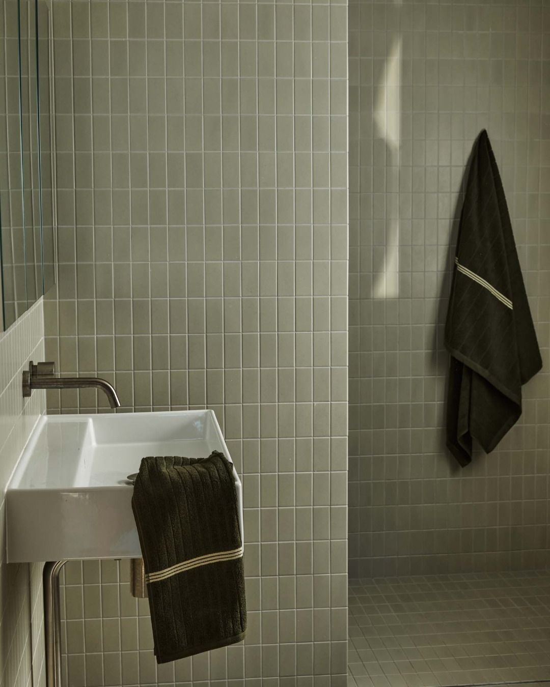 A pale green tiled bathroom with deep green hand towel and bath towels hung up