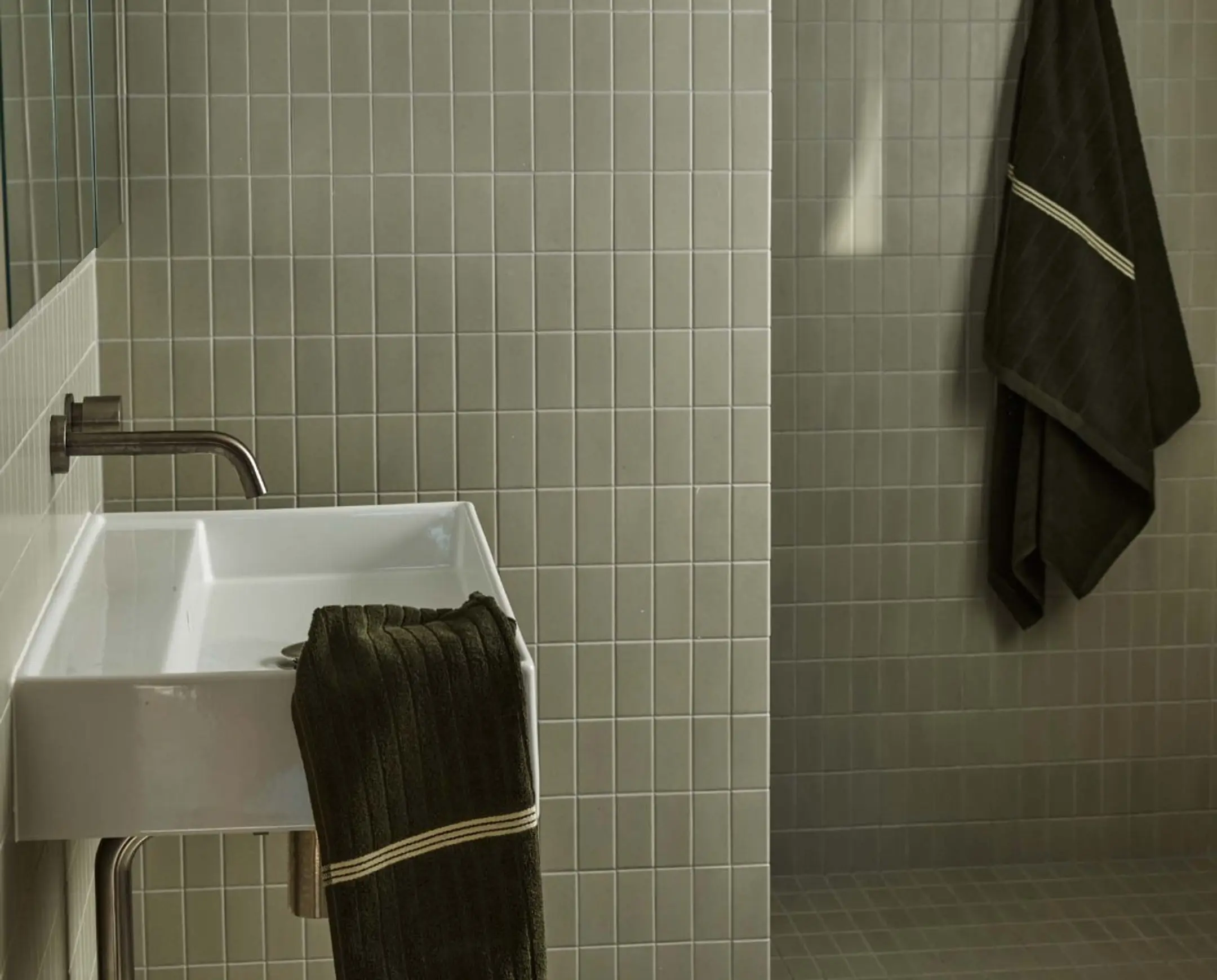A pale green tiled bathroom with deep green hand towel and bath towels hung up