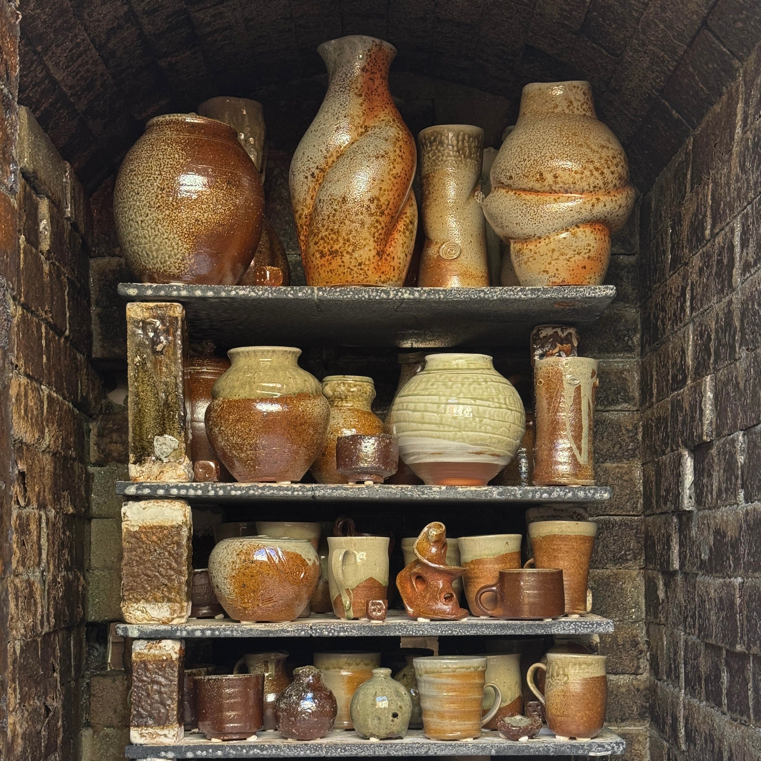 A collection of wood fired ceramics in the wood firing kiln stacked onto of each other in shelved units. Large forms of vases mixed with cups and mugs with natural rusty and burnished tones