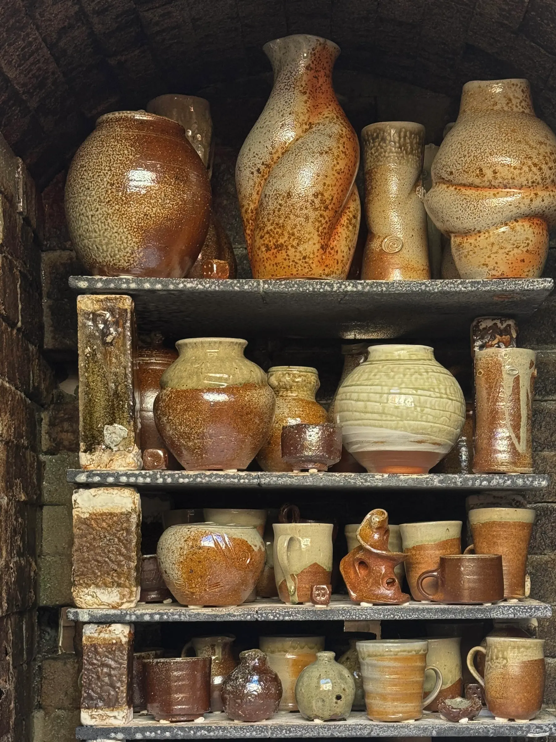 A collection of wood fired ceramics in the wood firing kiln stacked onto of each other in shelved units. Large forms of vases mixed with cups and mugs with natural rusty and burnished tones