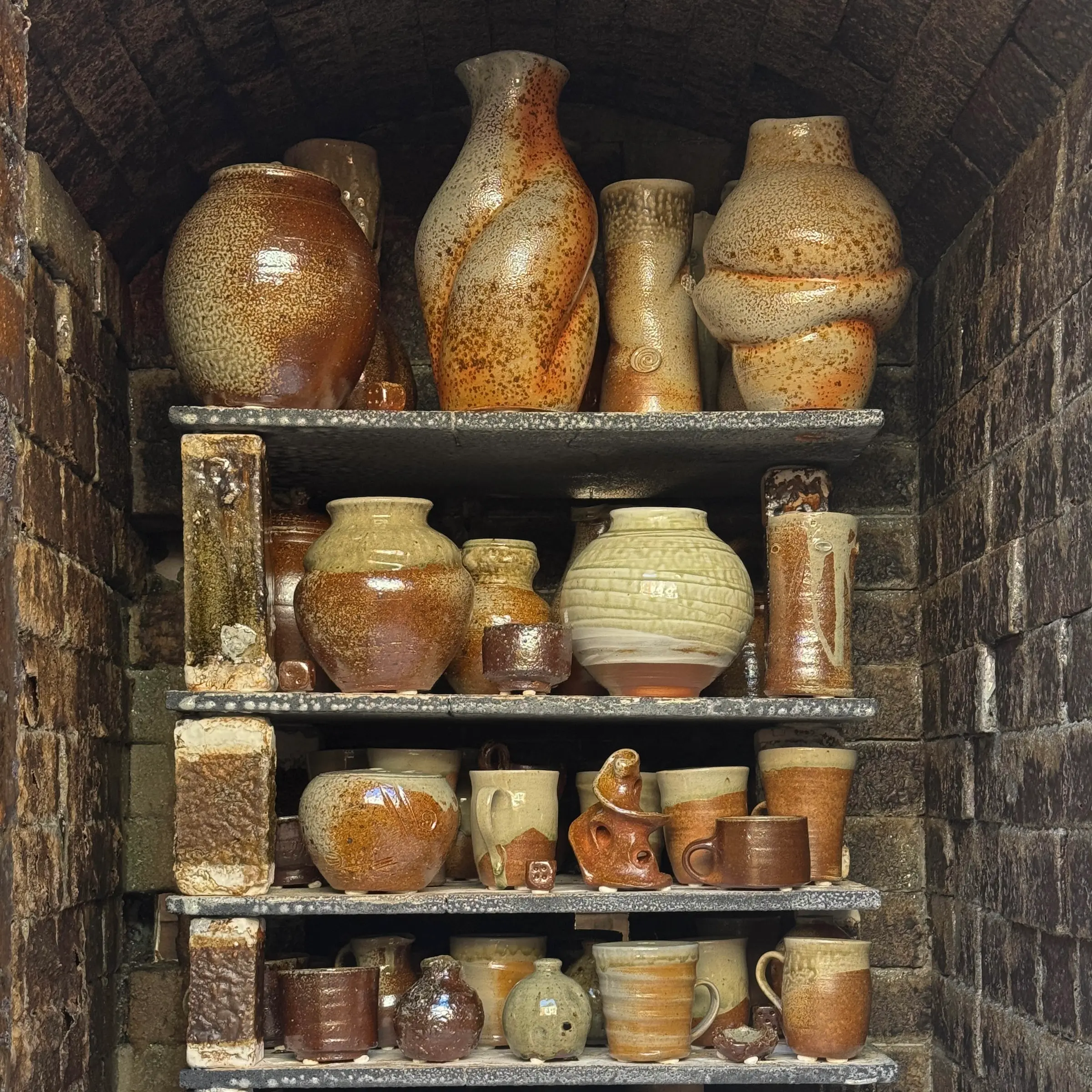 A collection of wood fired ceramics in the wood firing kiln stacked onto of each other in shelved units. Large forms of vases mixed with cups and mugs with natural rusty and burnished tones