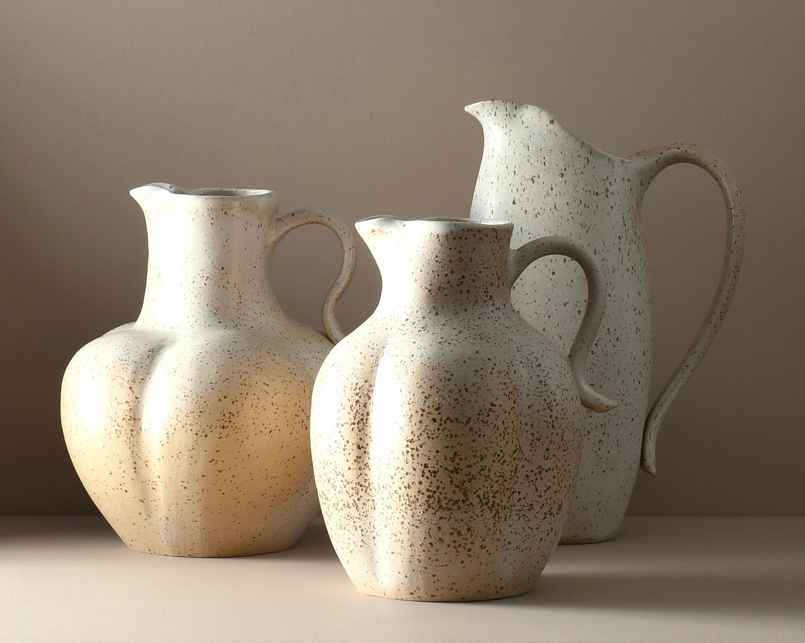 Three handmade ceramic jugs by Auckland artist Kaiqi Qiu, wheel-thrown and hand-altered in speckled cream stoneware with a soft satin eggshell glaze. Sculptural rounded forms with curved handles and pouring spouts, arranged in varying heights.