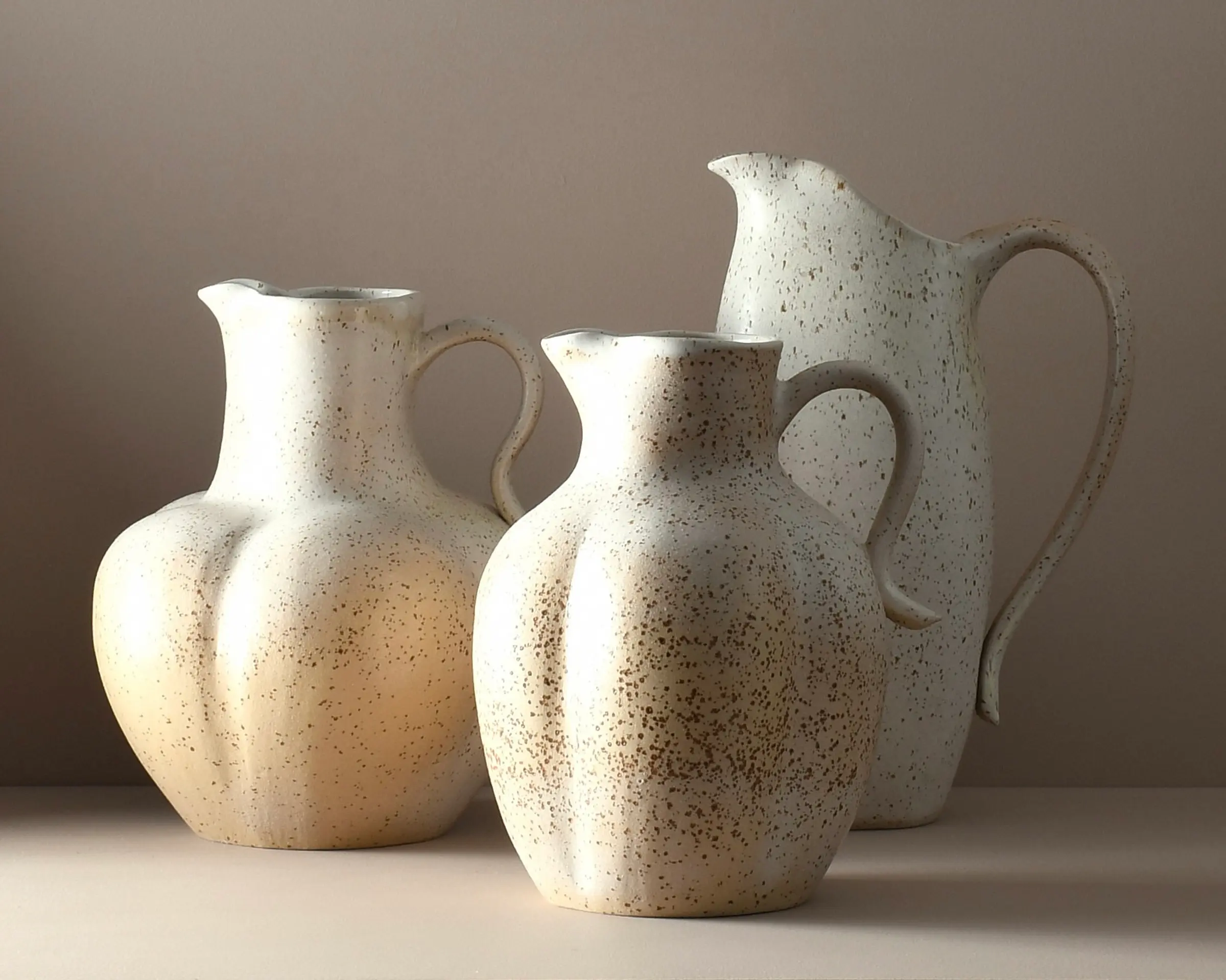 Three handmade ceramic jugs by Auckland artist Kaiqi Qiu, wheel-thrown and hand-altered in speckled cream stoneware with a soft satin eggshell glaze. Sculptural rounded forms with curved handles and pouring spouts, arranged in varying heights.