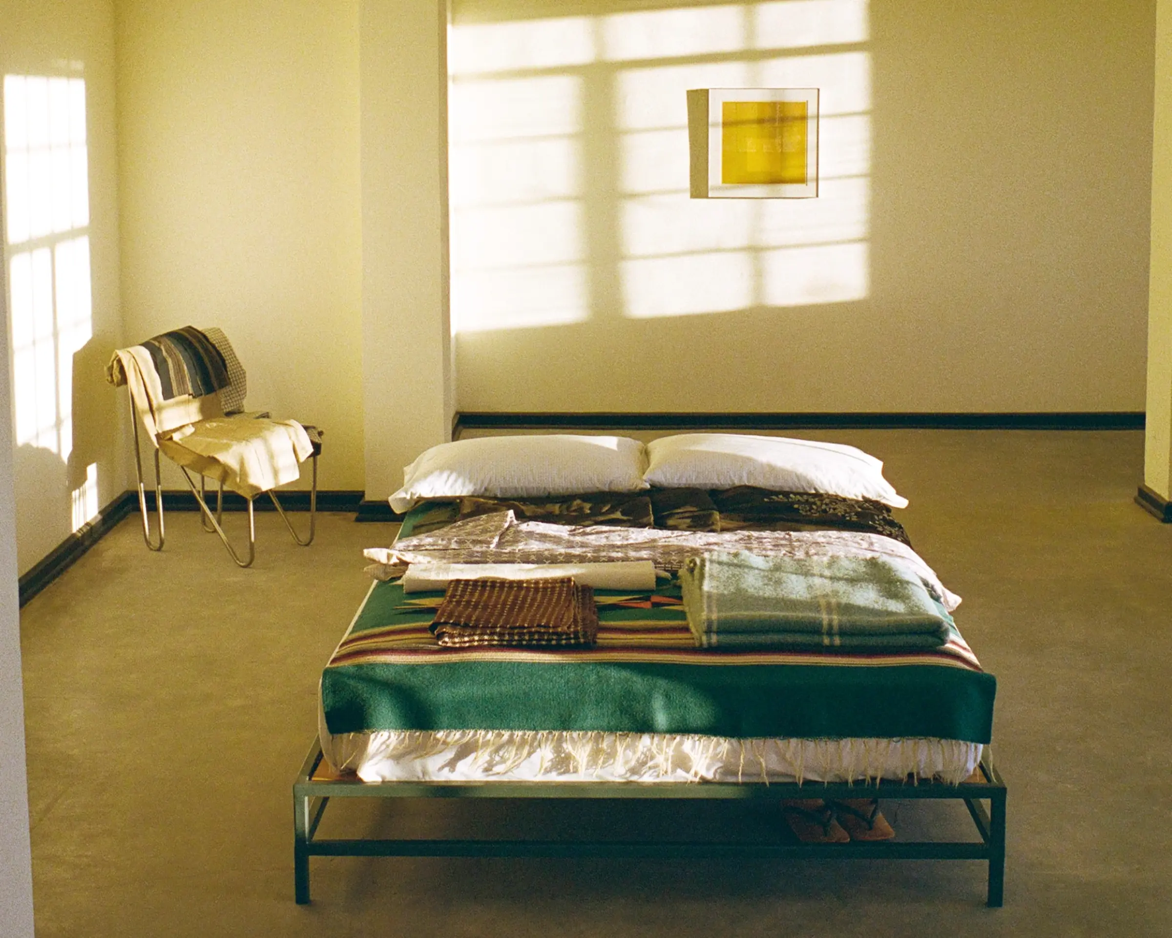 An image from Neptune Magazine Issue 10, a bed on a metal bed frame in a room with beige coloured walls, a singular painting with a chair in the corner and various textiles on the bed