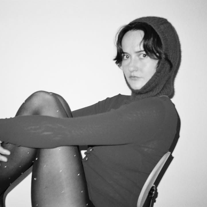 A black and white image of a woman wearing a Bonnie wore possum merino hand knit balaclava hat wearing a long sleep with tights on sitting on a chair