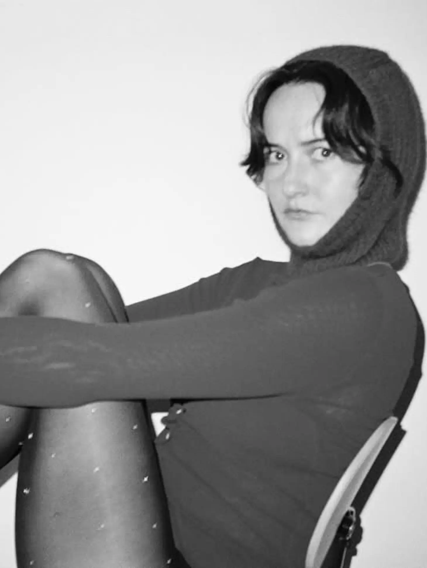 A black and white image of a woman wearing a Bonnie wore possum merino hand knit balaclava hat wearing a long sleep with tights on sitting on a chair