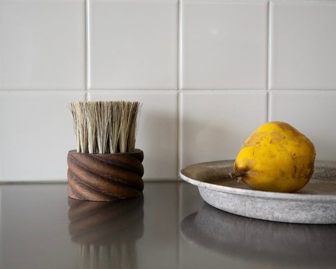 OROS Torsade vegetable brush in walnut-stained beechwood with natural horsehair and tampico fibres, photographed beside fresh fruit on a kitchen surface.