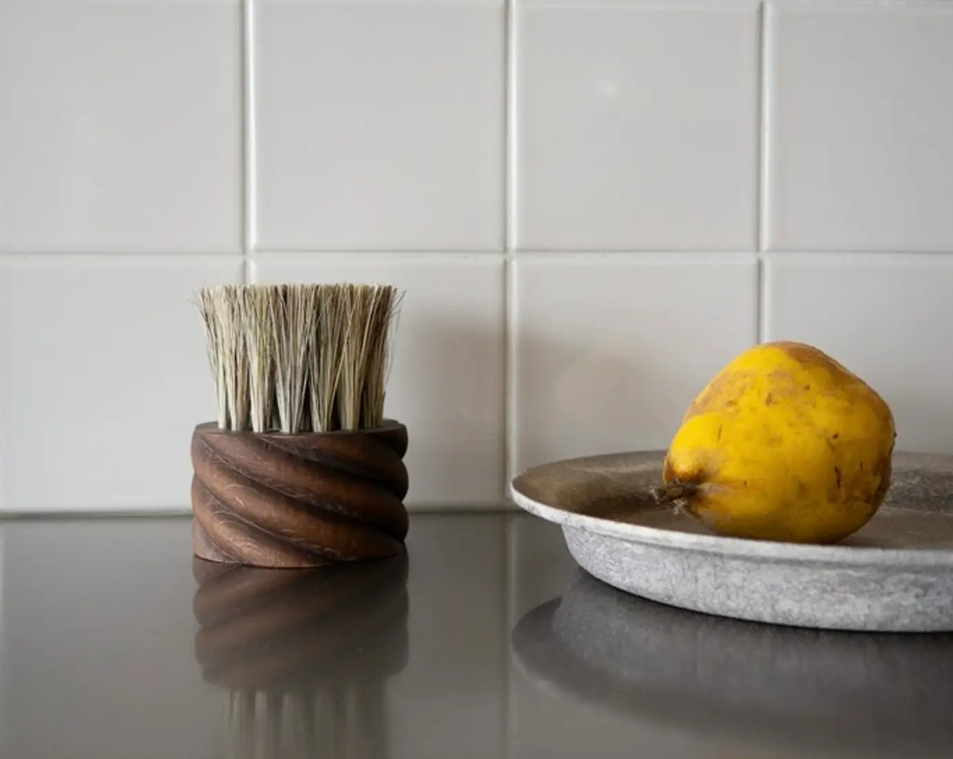 OROS Torsade vegetable brush in walnut-stained beechwood with natural horsehair and tampico fibres, photographed beside fresh fruit on a kitchen surface.