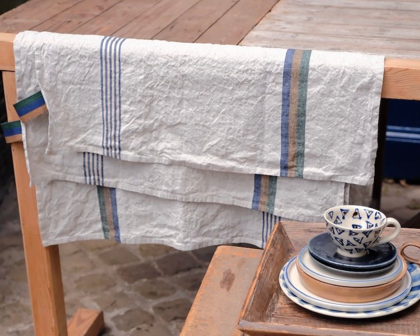 A libeco belgian striped tea towel hanging over a wooden railing with ceramic plates and a tea cup sitting infront