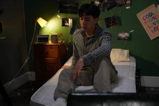 Leon in a stripe shirt sitting on a bed, depressed.