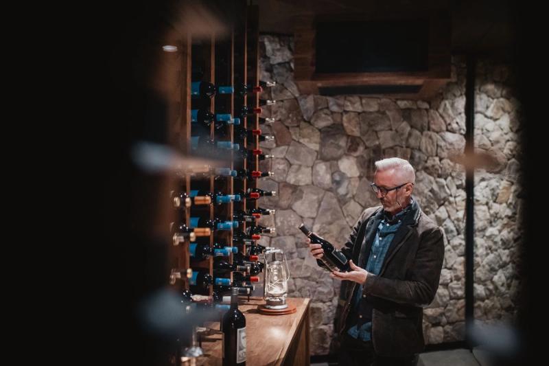 Sommelier with white hair and glasses examining a wine bottle in a stone-walled wine cellar with vertical wine racks