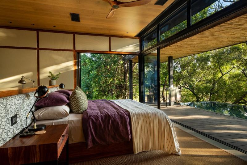 A modern bedroom with large glass walls opening to a wooden deck surrounded by lush green trees