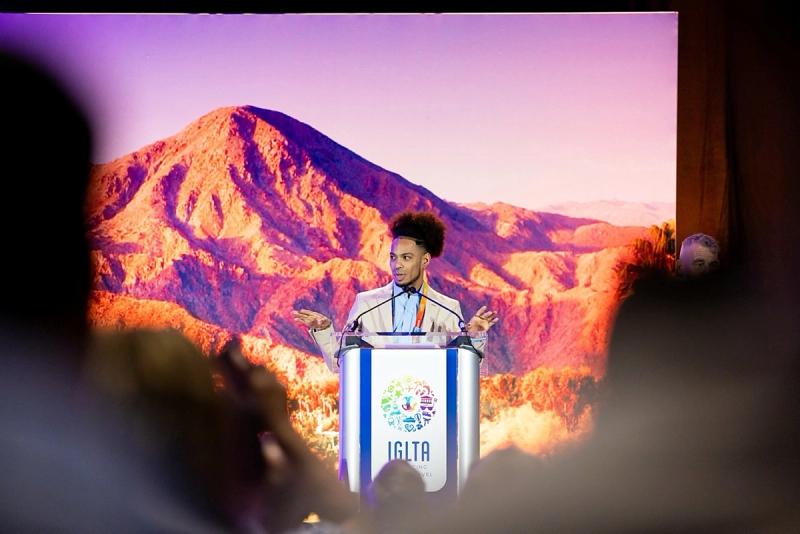 A speaker stands at a podium marked with the IGLTA logo, addressing an audience against a vivid mountain backdrop.