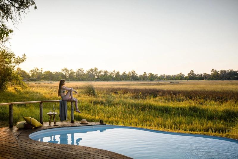 A wooden deck with a pool overlooks a golden savannah, where a woman sits on a railing, enjoying the peaceful view
