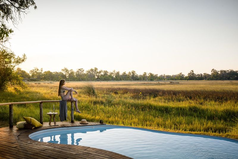 A wooden deck with a pool overlooks a golden savannah, where a woman sits on a railing, enjoying the peaceful view