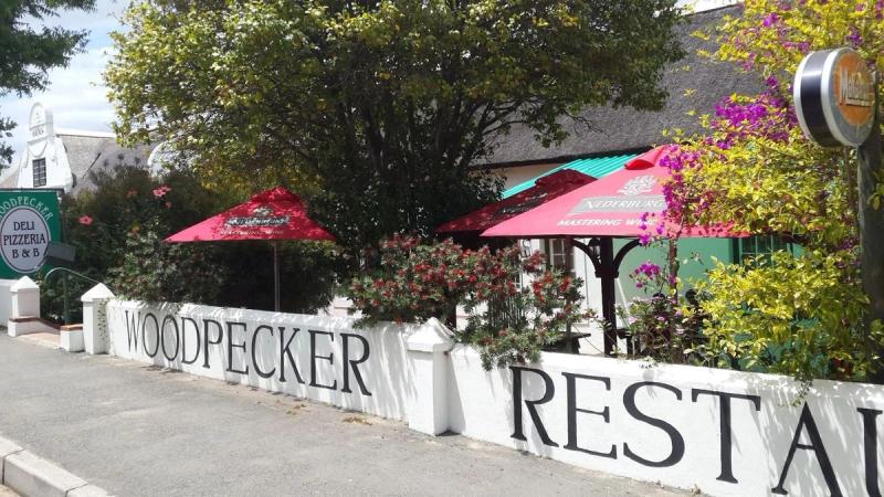 White wall with Woodpecker Restaurant signage beneath trees and red umbrellas in Swellendam